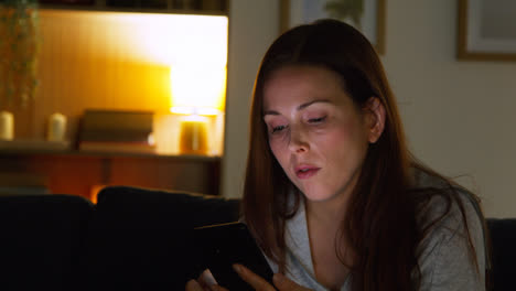 Mujer-Sentada-En-Un-Sofá-En-Casa-Por-La-Noche-Transmitiendo-O-Mirando-Contenido-En-Línea-En-Un-Teléfono-Móvil-1